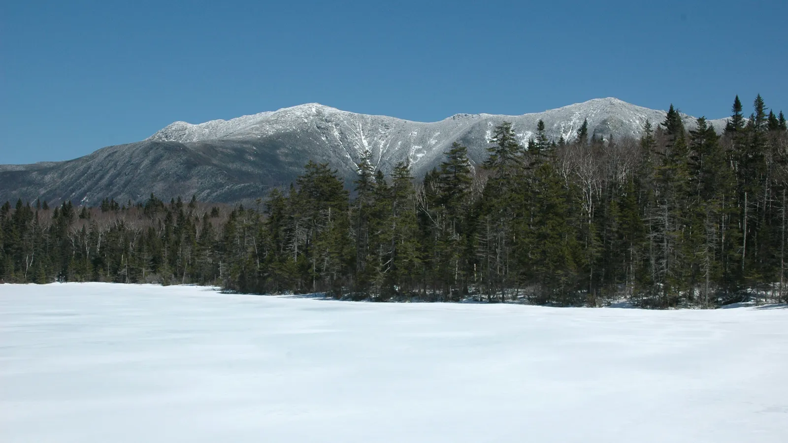 Snow-capped White Mountains ridge rising above evergreens and a frozen, snow-covered pond near Lincoln, New Hampshire