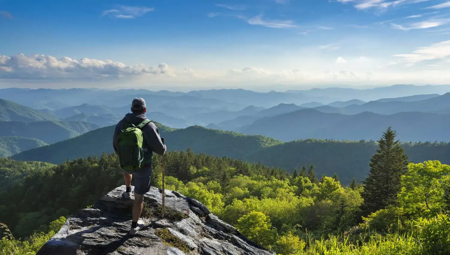 Hiker on an open rock ledge looking out over layered forested ridges of the White Mountains